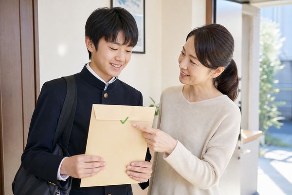 玄関で、学ラン姿の中学3年生の息子が持つ書類封筒のチェックマークを母親が指さし、提出前の確認をしている様子
