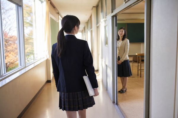 放課後の教室で、ノートを持った高校生が担当の先生へ相談しに行こうとしている場面