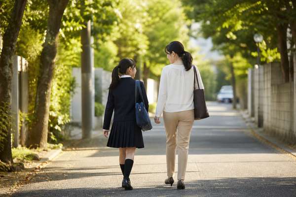 夕方の並木道を、制服の中学生と母親が並んで歩きながら話している後ろ姿