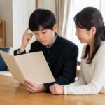 明るいリビングで学生服の中学生が通知表を開き、隣で母親が穏やかに見守っている様子
