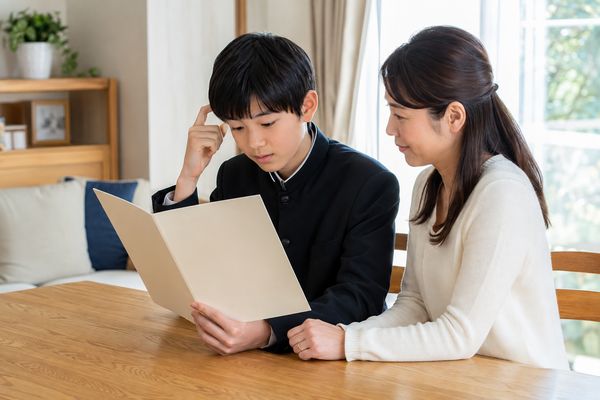 明るいリビングで学生服の中学生が通知表を開き、隣で母親が穏やかに見守っている様子