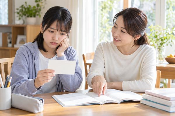 受験票を手に不安そうな表情の中学生と、英語の過去問を開いて指さす保護者