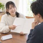自宅のテーブルで、高校生がノートの因数分解の式を指しながら講師に説明して笑顔になっている様子