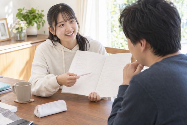 自宅のテーブルで、高校生がノートの因数分解の式を指しながら講師に説明して笑顔になっている様子