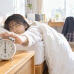 朝の寝室で中学生が布団から手を伸ばして6時30分を指す目覚まし時計を止めようとしている様子