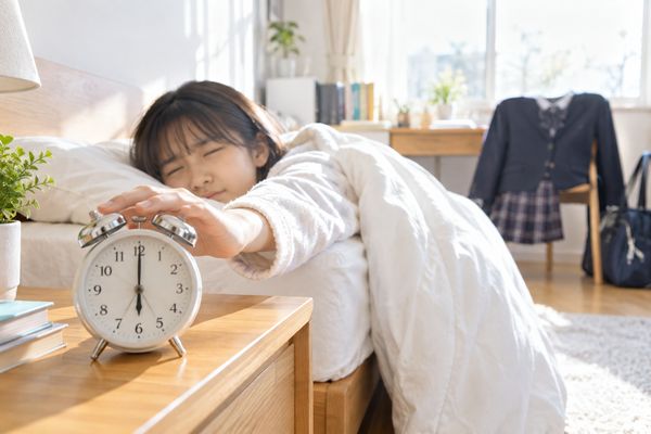 朝の寝室で中学生が布団から手を伸ばして6時30分を指す目覚まし時計を止めようとしている様子
