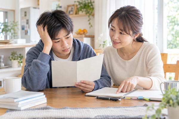 自宅のダイニングで、オール2の通知表を見て困った表情の中学生に保護者が寄り添って話している様子