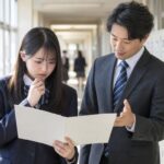 明るい高校の廊下で、通知表を開いて不安そうな高校生に先生が評価の見方を説明している様子