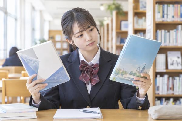 図書館で2冊の英語の教材を見比べ、どちらを使うか考える制服姿の高校生