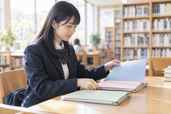 制服姿の高校生が図書館で色の違う三つの書類ファイルを見分けている様子