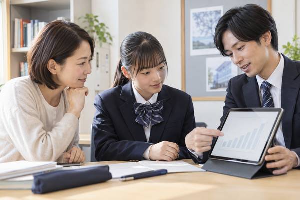 塾の面談席で、保護者と制服姿の生徒が、塾スタッフの示すタブレットの成績推移グラフを一緒に見ている様子