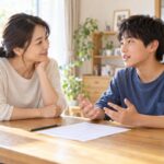 自宅のダイニングで、答案用紙を前に中学生の息子が話し、母親が頬杖をついて耳を傾けている様子