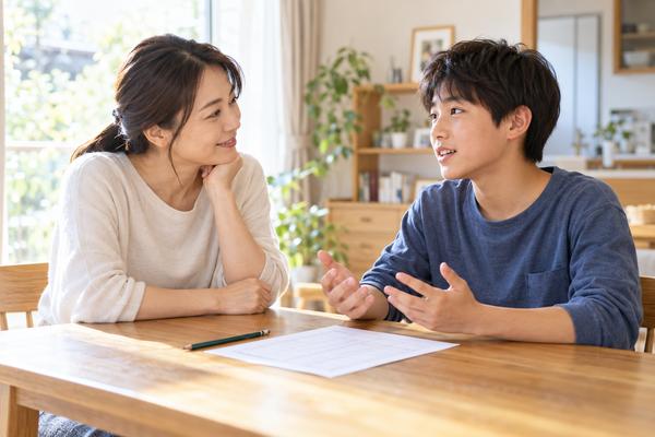 自宅のダイニングで、答案用紙を前に中学生の息子が話し、母親が頬杖をついて耳を傾けている様子