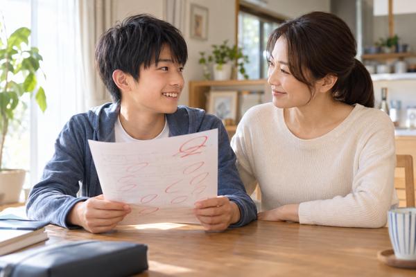 自宅のテーブルで、赤ペンの入った答案を手にした中学生と母親が、笑顔で向き合って話し合っている様子