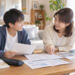 リビングのテーブルで、中学生の息子と母親が高校の学校案内や模試の成績表を広げ、笑顔で志望校について話し合っている様子