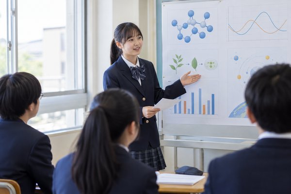 明るい教室で、制服姿の生徒が自作のポスターを指しながらクラスメイトの前で発表している様子