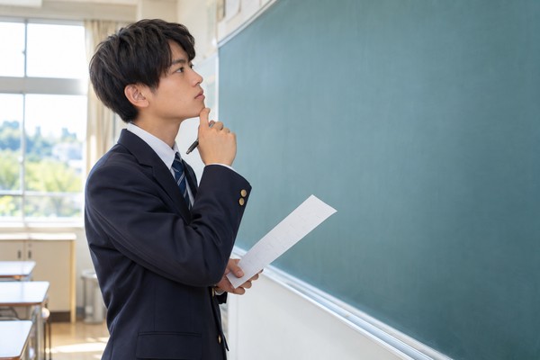 制服姿の高校生が教室の黒板の前で問題用紙を手に、最初の一手を考え込んでいる様子