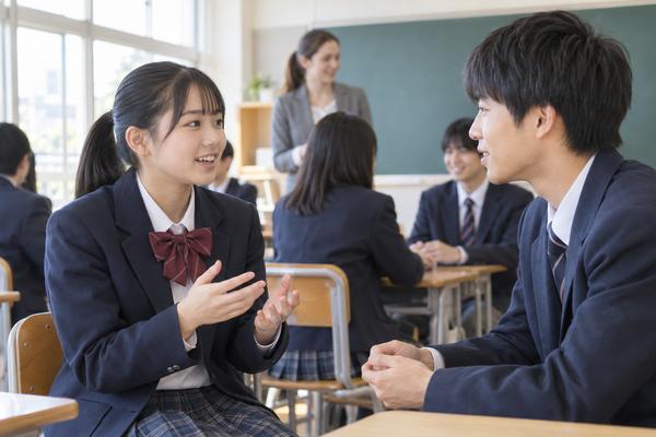 明るい教室で制服姿の中学生が二人で向かい合い、英語で会話のやり取りを練習している様子