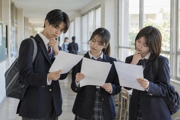 校舎の廊下で制服姿の高校生3人が返ってきた答案を見せ合い、点数を比べている様子