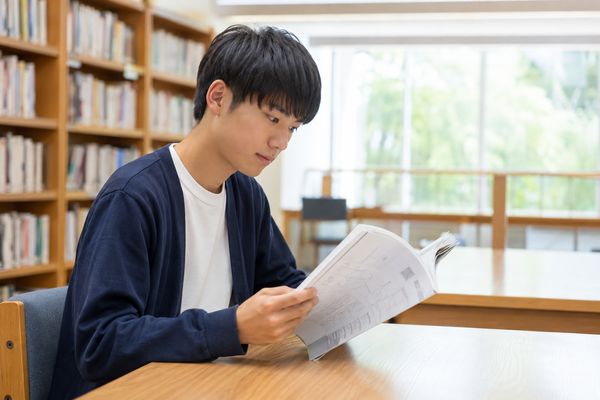 明るい図書館の閲覧席で、私服の男子高校生が数学の月刊誌のページをめくって内容を確かめている様子
