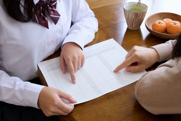 制服の中学生と保護者が食卓で通知表風の用紙を広げ、二人の指がそれぞれ別の評定欄を指している手元