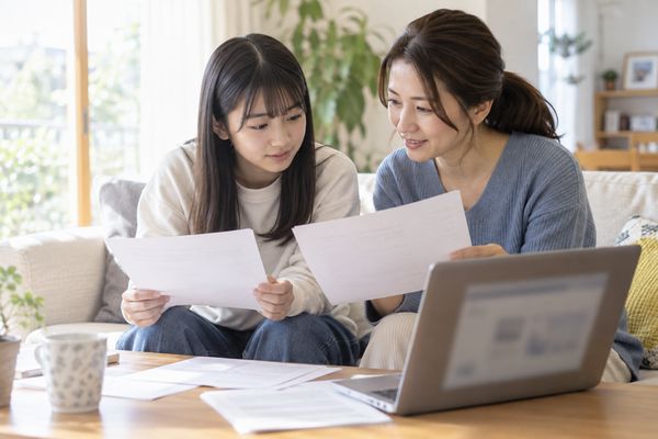 明るいリビングで高校生と母親が慶應の学部別の入試資料を見比べている様子
