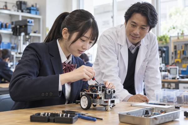 理科室でロボットを組み立てる高校生と、白衣で見守る先生
