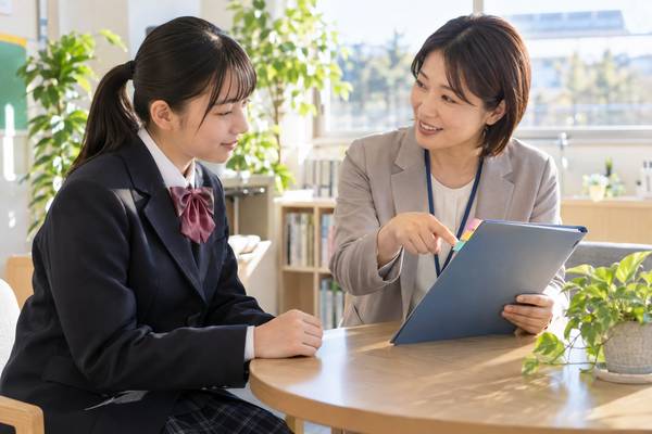 放課後の相談スペースで、女性の先生が3色の付箋を貼った通知表フォルダを指しながら制服姿の生徒に説明している場面