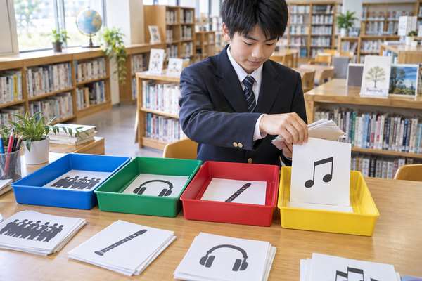 学校の図書室で、制服の中学生が歌唱・鑑賞・器楽・楽典を表す4色のトレーに資料を仕分けている様子