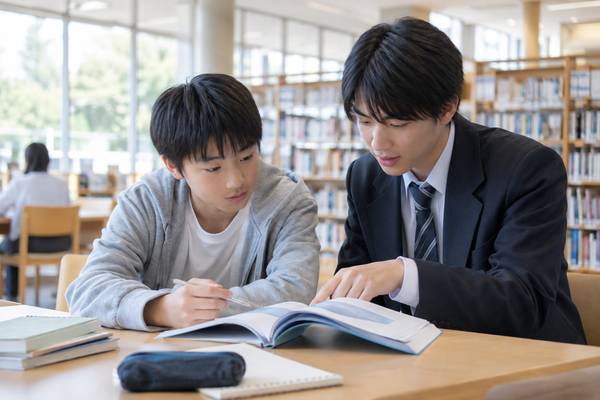 明るい図書館で、私服の中学生が高校生の先輩といっしょに問題冊子をのぞき込んでいる様子。中学生が過去問に挑戦して高校内容の理解度を確かめている場面