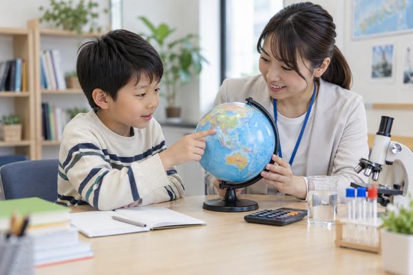 明るい学習塾で地球儀を指さしながら講師と話す小学生と、机に置かれた理科の実験道具