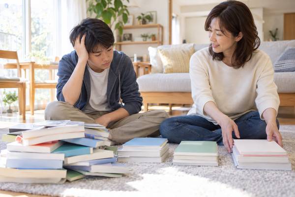 リビングの床に何冊もの参考書を積み上げ、どれから手をつければよいのか分からず頭を抱える中学生と、隣で見守る母親