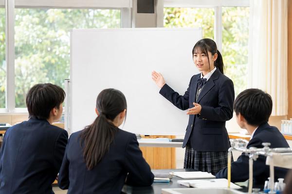 理科室のホワイトボードの前で、制服姿の女子高校生が身ぶりを交えて考えの根拠を説明し、同級生が聞いている様子