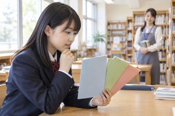 図書室で、制服姿の中学生が色違いの3冊の資料を並べて見比べている場面