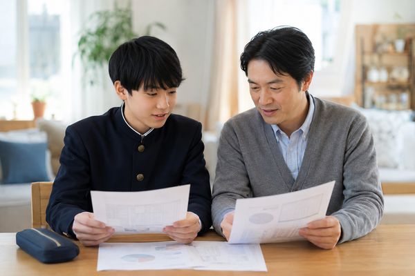 学生服の中学生と父親がダイニングテーブルで通知表と模試の成績表を並べて読んでいる様子