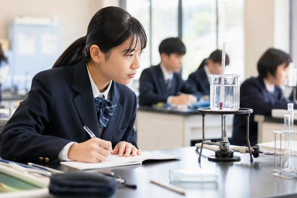 明るい理科室で制服の中学生が実験器具を観察しながら、気づいたことをノートに書き留めている様子