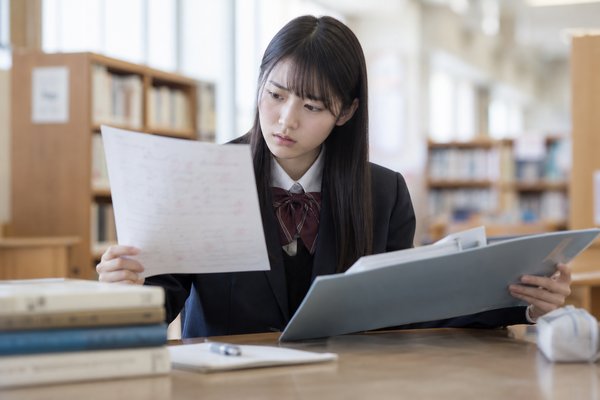 学校の図書室で、制服の高校生が採点された答案と提出用ファイルを見比べて納得できない表情をしている様子
