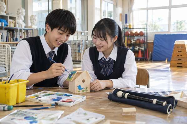 中学校の明るい教室で木工作品に色を塗る制服の中学生二人と、机に並んだリコーダーや絵の具