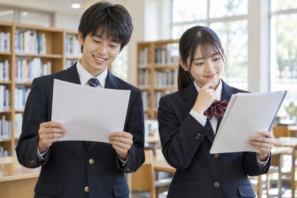 明るい学校の図書室で、制服姿の中学生2人がそれぞれ別の学習成果物を持って見比べている様子