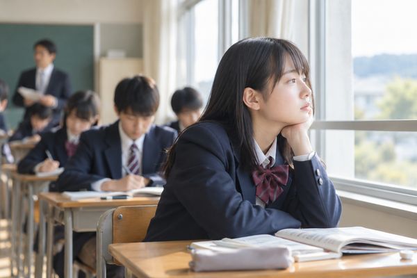 明るい教室で、周りの生徒がノートに書き込むなか、ひとり頬杖をついて窓の外へ視線を向けている制服姿の生徒
