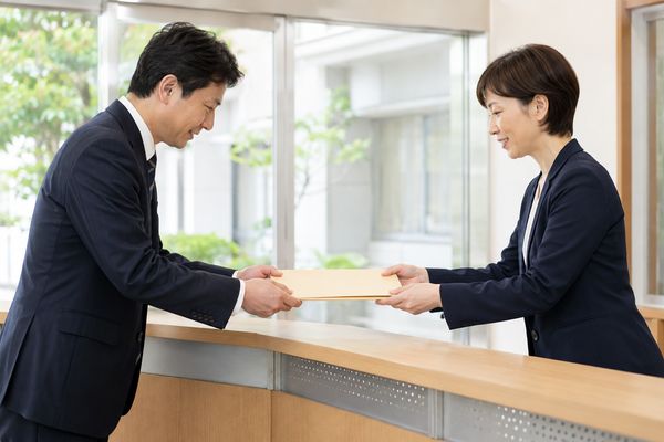 中学校の職員が高校の受付で、指導要録の写しが入った書類フォルダーを高校の職員へ手渡している様子