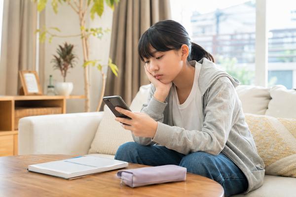 リビングのソファでスマートフォンを見つめ、教材を閉じたままにしている中学生