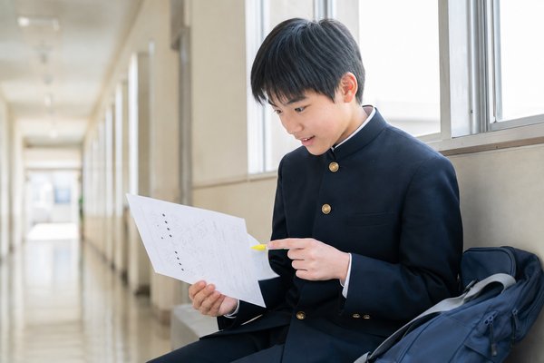 明るい校舎の廊下で、学ラン姿の中学生が返却された英語の問題用紙に蛍光ペンで印を付けながら見返している様子