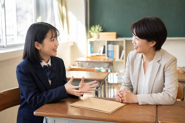 放課後の明るい教室で担任の先生に活動の様子を笑顔で話す女子中学生
