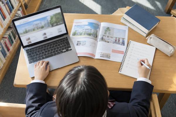図書室でノートパソコンの大学入試ページと募集要項を見ながらノートに書き写す高校生