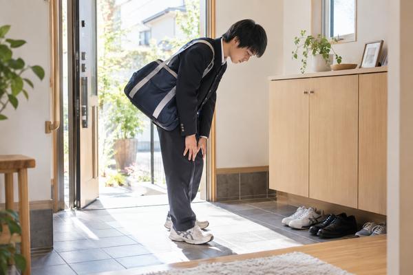 朝の玄関で、学ランに部活バッグを肩掛けした中学生の男子が疲れた様子でうつむき、立ち止まっている様子