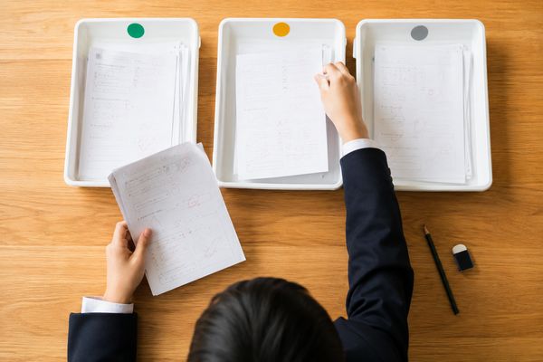 制服姿の中学生が返却答案を緑・黄・グレーの丸印が付いた3つのトレーに仕分けている手元の俯瞰