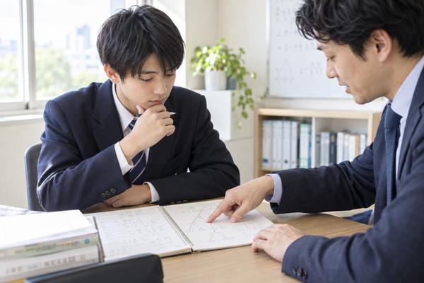 制服姿の高校生の隣で、講師がノートに描いた枝分かれの図の一点を指し、方針の分岐点を振り返っている様子