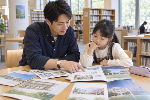 明るい図書館で父親と小学生が複数の学校案内を机に並べて見比べている様子