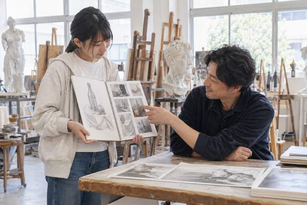 アトリエでデッサンを手にした美大志望の生徒が、講師から具体的な指導を受けている様子